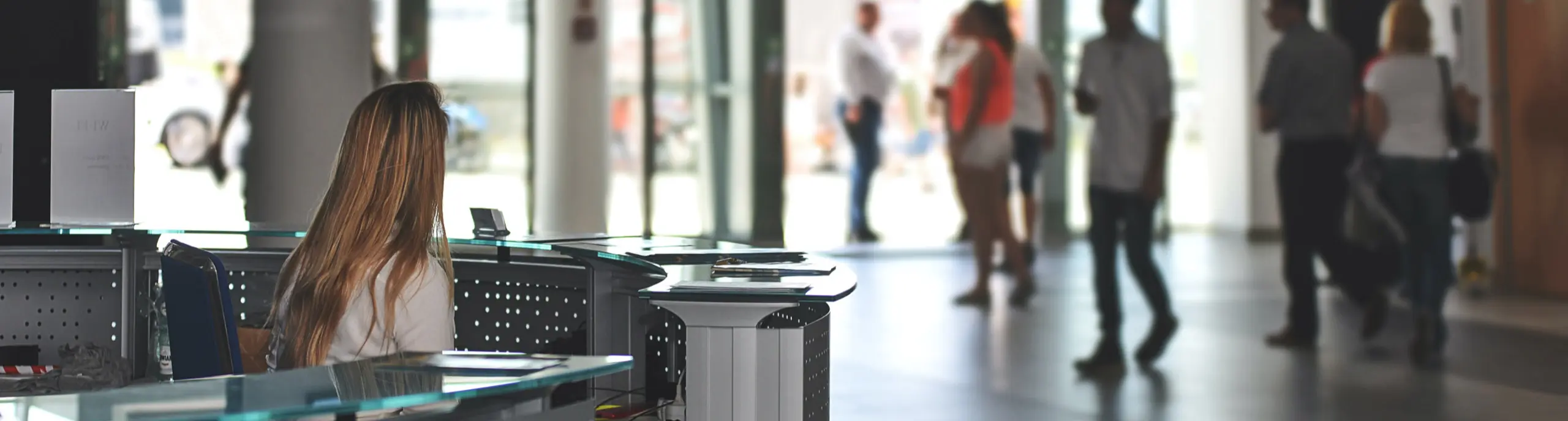 A reception desk with a female receptionist and blurred figures of people walking in the background